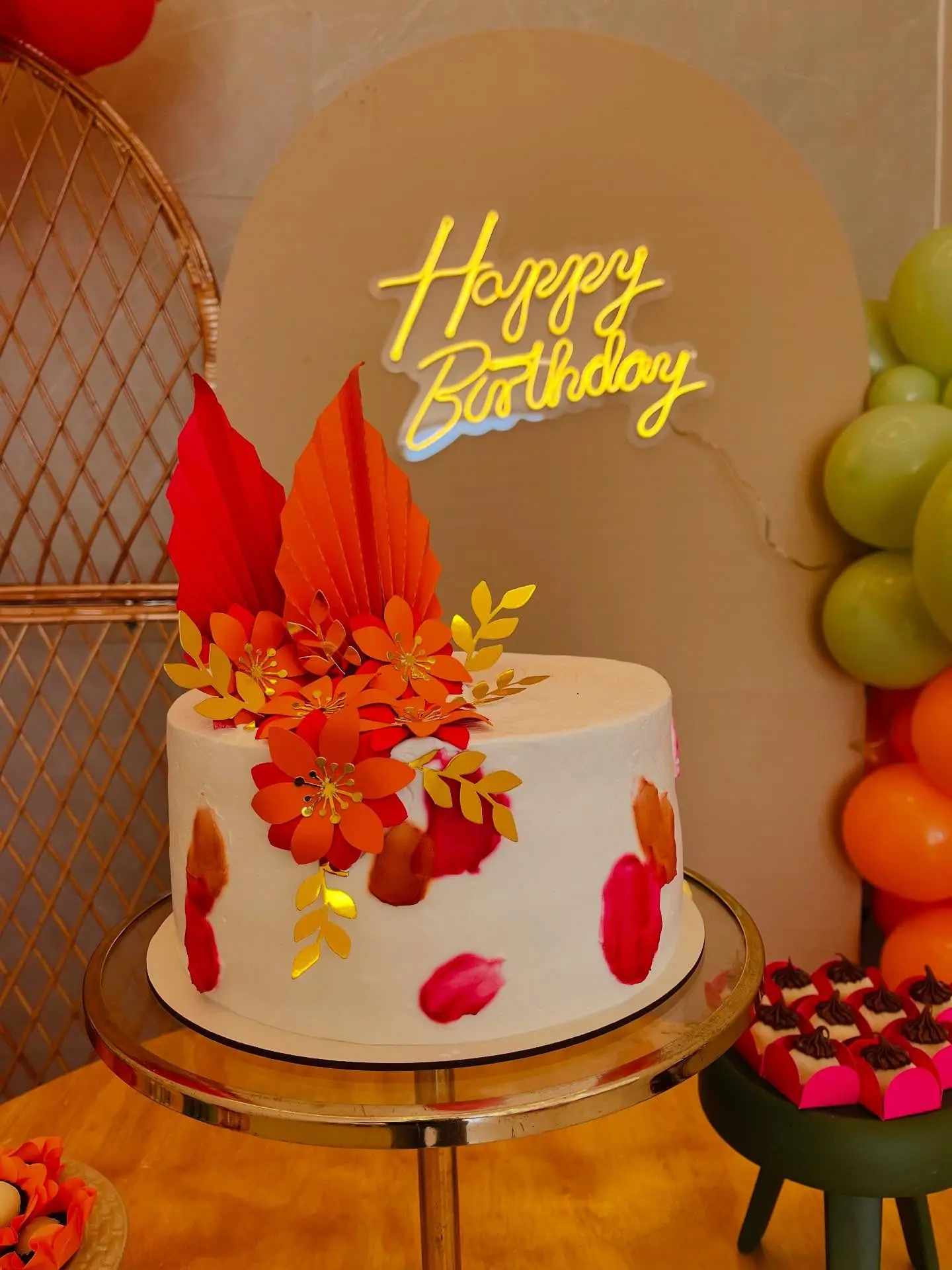 Bolo de aniversário branco com folhas e flores em tons laranja diante de painel Happy Birthday